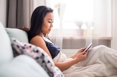 Kanepede oturan genç Asyalı kadın okuyucu tableti kullanıyor ve evde kitap okuyor.