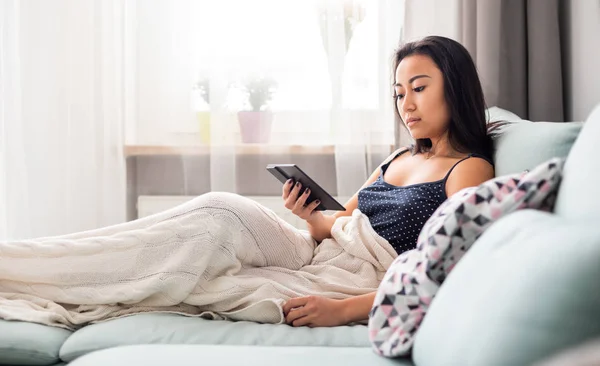 Joven mujer asiática sentada en un sofá usando una tableta lectora y leyendo un libro electrónico en casa —  Fotos de Stock