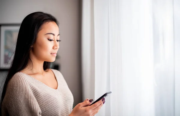 Giovane donna asiatica utilizzando smartphone a casa in piedi vicino alla finestra — Foto Stock