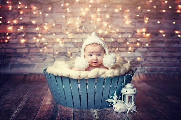 Lovely, beautiful newborn boy lying in blue basket in a white hat, new year and Christmas tree decorations, garland on the wall, beautiful bokeh, with a flashlight. card, text. toning. winning smile — Stock Photo, Image