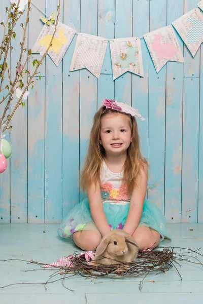 Little beautiful and charming girl sister along with rabbit sitting on the floor or chair in bright blue clothes Easter, eggs, festive mood, emotion and smile surprise holiday celebration — Stock Photo, Image