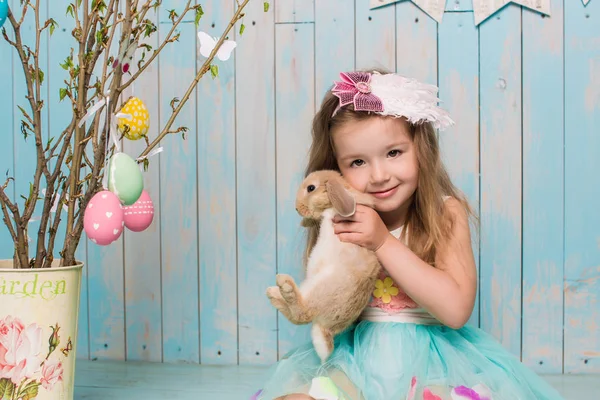 Little beautiful and charming girl sister along with rabbit sitting on the floor or chair in bright blue clothes Easter, eggs, festive mood, emotion and smile surprise holiday celebration — Stock Photo, Image