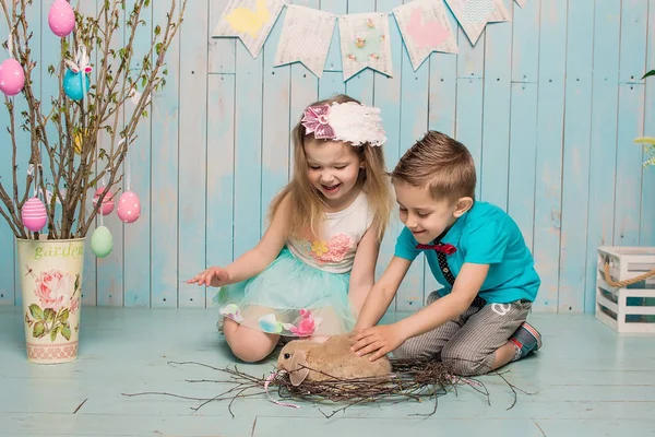 Two little childs, boy, girl, brother and sister along with rabbit sitting on the floor in bright blue clothes Easter, eggs, festive mood, emotion and smile surprise holiday celebration — Stock Photo, Image