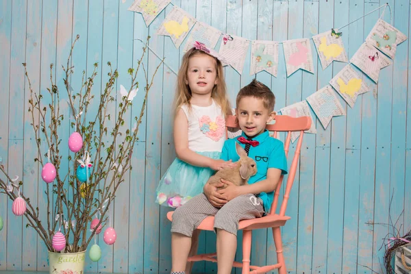 Two little childs, boy, girl, brother and sister along with rabbit sitting on the floor in bright blue clothes Easter, eggs, festive mood, emotion and smile surprise holiday celebration — Stock Photo, Image