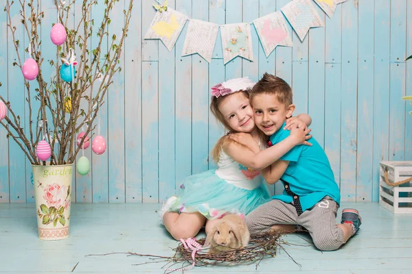 Två små childs, pojke, flicka, bror och syster tillsammans med kaninen sitter på golvet i ljusa blå kläder påsk, ägg, festlig stämning, känslor och leende överraskning semester firande Royaltyfria Stockfoton