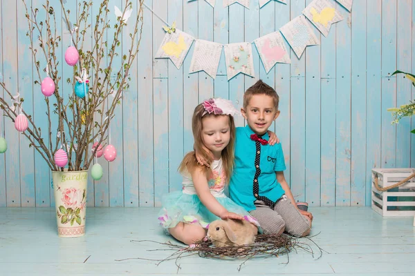 Twee kleine childs, jongen, meisje, broer en zus samen met konijn zittend op de vloer in helder blauwe kleding Pasen, eieren, feestelijke stemming, emotie en glimlach verrassing vakantie feest Stockfoto