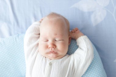 Üç aylık bebek yastıkta uyuyor. Gün ışığında kolları havada.