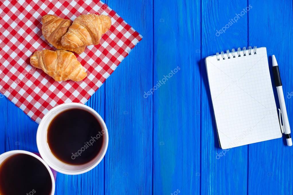 Two cups of coffee, croissants and open notepad with pen on blue