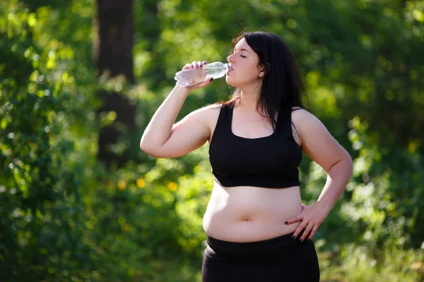 Young thirsty overweight woman drinking water after jogging in t — Stock Photo, Image