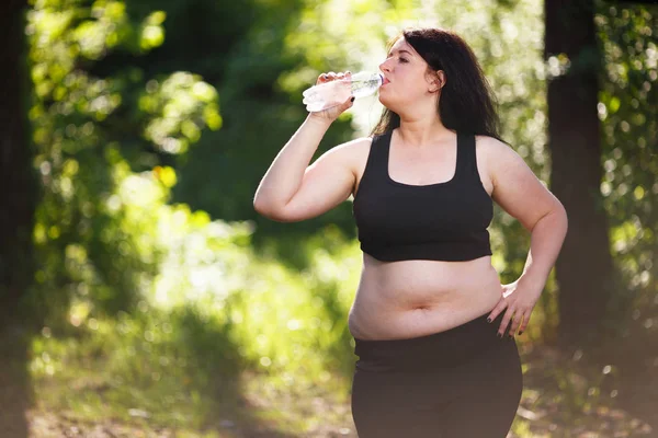 Young thirsty overweight woman drinking water after jogging in t — Stock Photo, Image