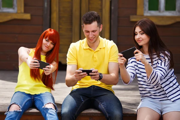 Three young friends sitting on bench addicted to mobile video ga