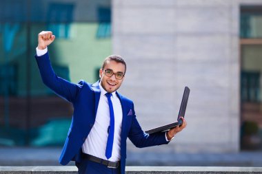 Kol kaldırdı tutmak dizüstü bilgisayar ile genç işadamı ve açık havada arka planda bina ofis ile ayakta iken pozitifliği ifade heyecan. Başarı kutluyor