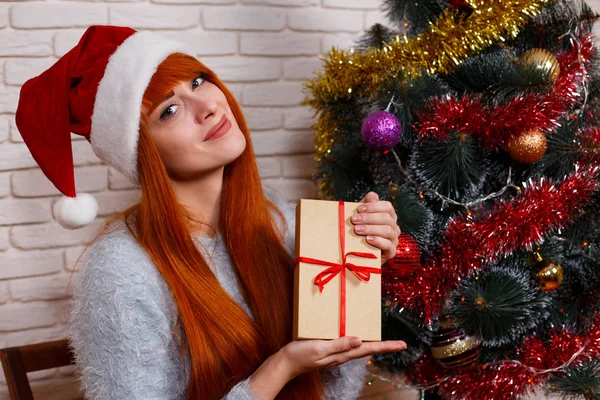 Beautiful young girl in Santa cap holding a gift box sitting nea — Stock Photo, Image