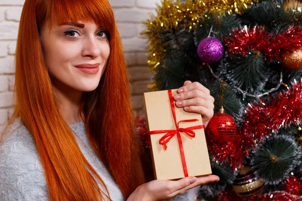 Beautiful young redhead girl holding a gift box sitting near Chr — Stock Photo, Image