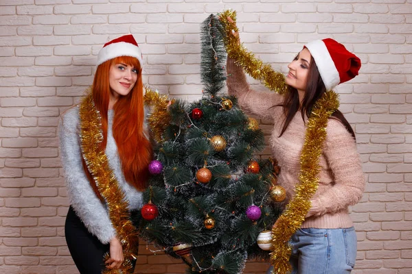 Two young beautiful friends in Santa caps decorating Christmas t — Stock Photo, Image