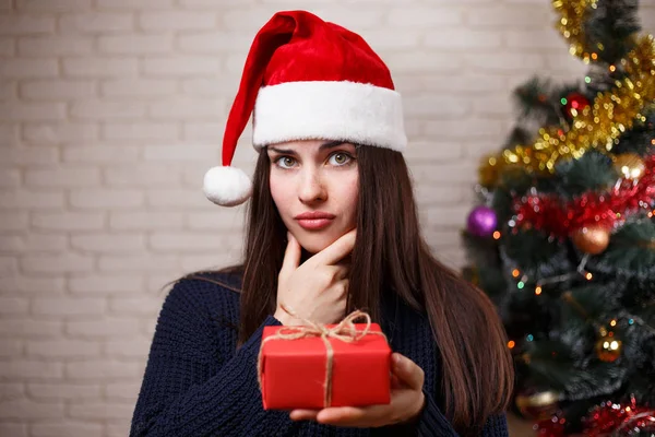 Young cute pensive woman in Santa cap with a gift box in hand. N — Stock Photo, Image