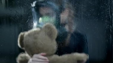Endişeli çocuk oyuncak ayıyı sıkıca tutarken gaz maskesi takıyor. Hava karanlık ve pencereden yağmur yağıyor.. 