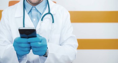 Beyaz erkek doktor laboratuvar önlüğü ve steteskoplu eldivenler giyiyor cep telefonuyla iş arkadaşlarıyla iletişim kuruyor..