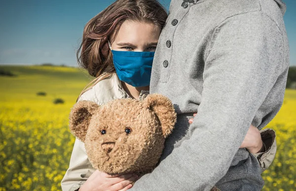 Caucasian Girl Wearing Blue Safe Breathing Mask Hugging Her Dad and Teddy Bear During Pandemia. Scenic Sunny Landscape in Background. Happy Family Time.