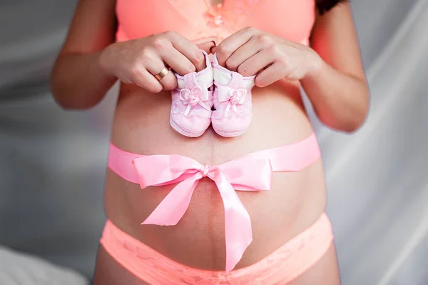 O fată însărcinată cu un arc pe stomac ține șosete pentru copii . — Fotografie, imagine de stoc