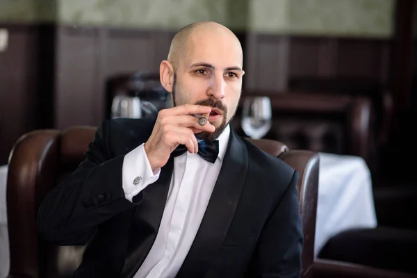 A brutal man in a suit smokes a cigar.