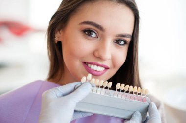 Dişçi genç kadın dişlerinin rengini kontrol ediyor ve seçiyor.