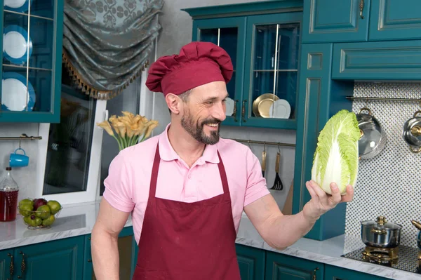 Very happy man holding fresh chinese cabbage. Cook with big smile and pakchoi in hand. Chef talks to vegetables. Portrait of happy cook in hat on kitchen. Guy in red apron prepared vegetables to eat