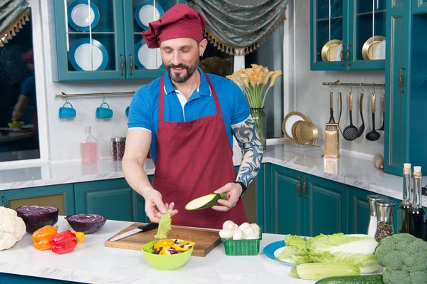 Tattooed chef in apron and cap cooking salad. Guy cooking vegetable salad with red cabbage and zucchini in yellow plate. Cook shows how to make vegetable salad. tattooed man loves to work on kitchen.