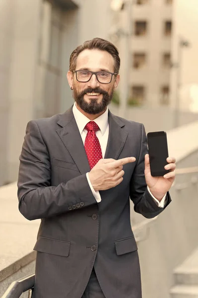 Businessman and digital gadget. Portrait of smiling businessman pointing finger at blank screen mobile phone isolated over building background. Happy businessman pointing at smartphone in hand.