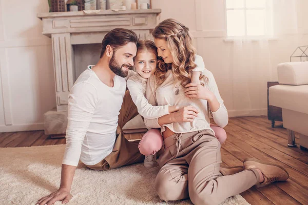 Gelukkig jong gezin met een kind zittend op een tapijt en knuffelen thuis — Stockfoto