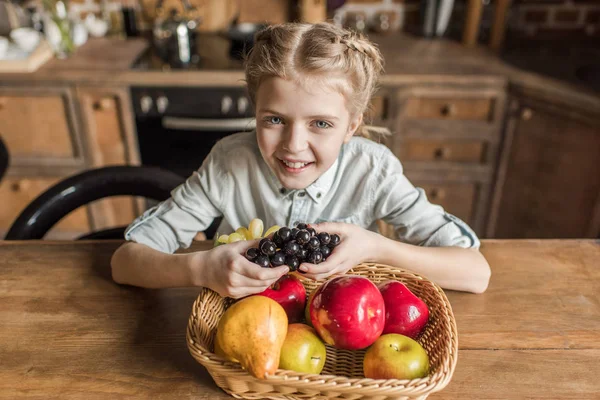 Adorable girl with different fruits sitting at table in kitchen — Stock Photo, Image