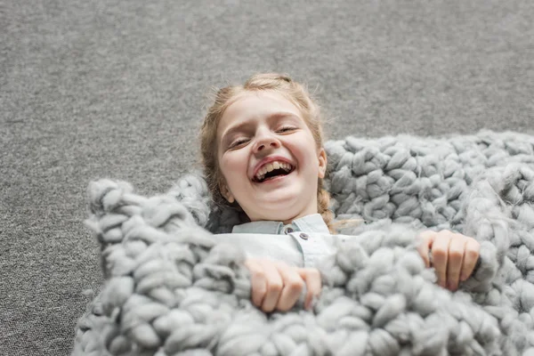 Adorable girl laughing and lying on floor with grey knitted blanket — Stock Photo, Image