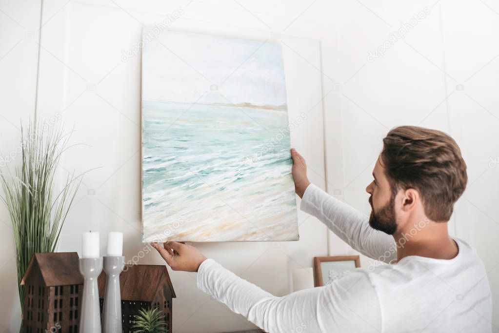 Young bearded man hanging picture on wall at home