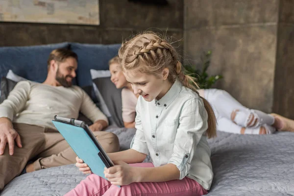 Urocza córka, za pomocą cyfrowego tabletu i leżąc na łóżku z rodzicami — Zdjęcie stockowe