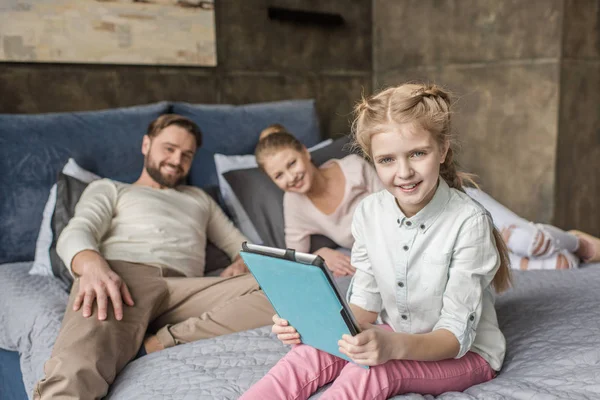 Schattige dochter met behulp van digitale tablet en liggend op bed met ouders — Gratis stockfoto