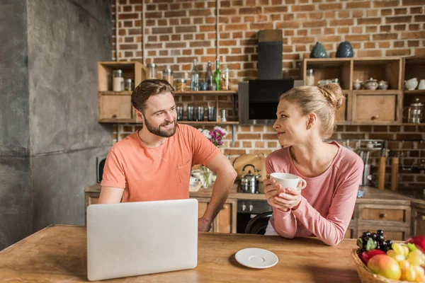 Feliz pareja joven utilizando el ordenador portátil en la cocina y sonriendo entre sí —  Fotos de Stock