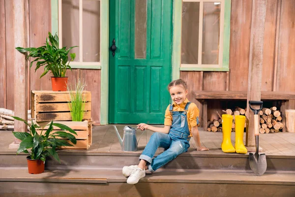 Adorable smiling little girl sitting on porch with watering can — Stock Photo, Image