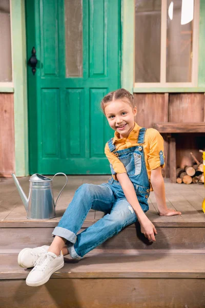 Adorable smiling little girl sitting on porch with watering can — Stock Photo, Image