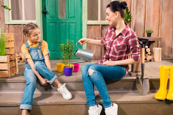 Side view of smiling mother and daughter sitting on porch and watering plant — Stock Photo, Image