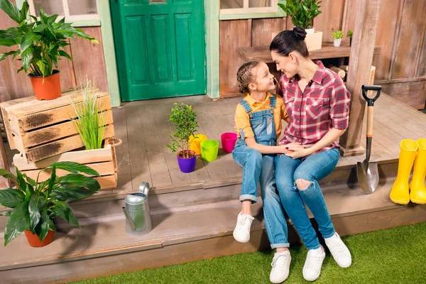 Happy mother and daughter sitting and holding hands on porch with potted plants — Stock Photo, Image