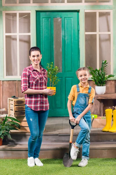 Усміхнена мати з горшковою рослиною і дочкою з садовим лопатою дивиться на камеру — стокове фото