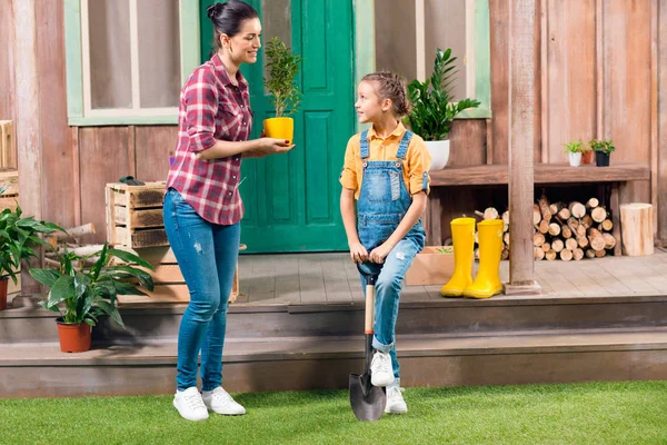 Madre sonriente con planta en maceta e hija con pala de jardín mirándose entre sí — Foto de Stock