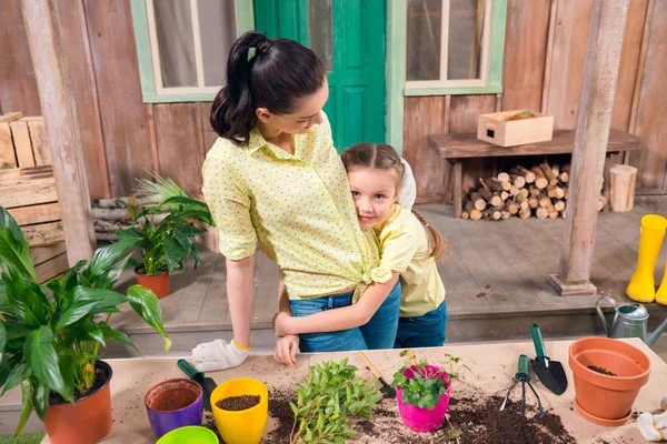 Mère et fille avec des plantes et des pots de fleurs debout et étreignant à table sur le porche — Photo