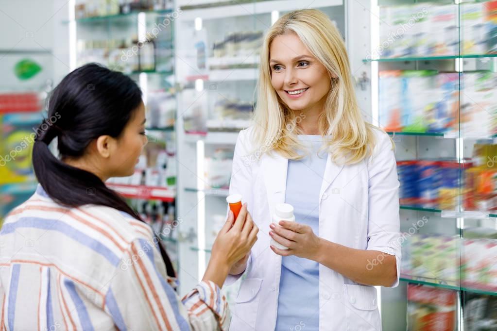 Smiling female pharmacist showing containers with pills to customer in drugstore
