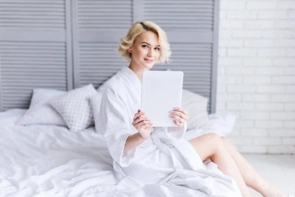 Beautiful Blonde Girl Bathrobe Holding Digital Tablet Smiling Camera Bedroom — Free Stock Photo