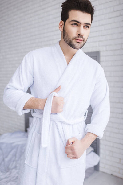 handsome young man tying belt of bathrobe and looking away at home