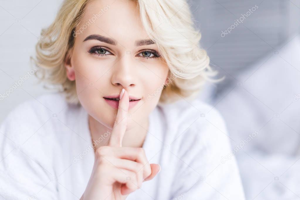 beautiful blonde woman in bathrobe gesturing for silence and looking at camera