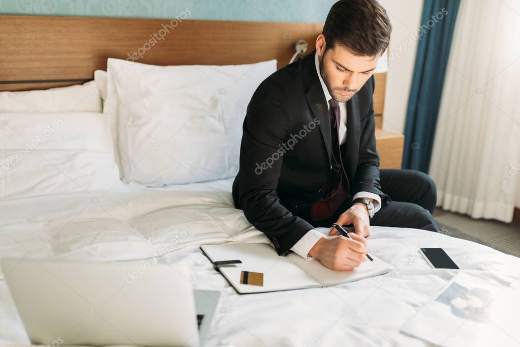 businessman writing something to notebook while sitting on bed in hotel room