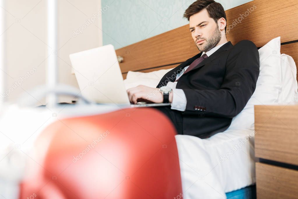 businessman lying on bed in hotel room and using laptop 