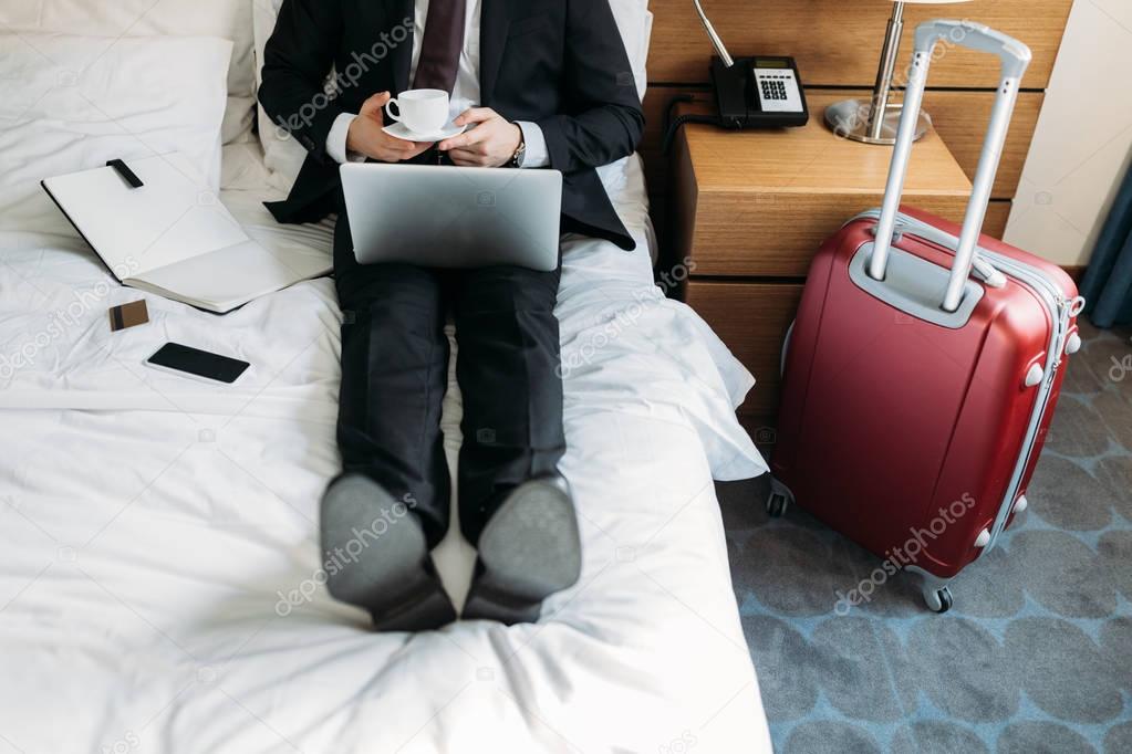 cropped image of businessman lying on bed in hotel with coffee and laptop